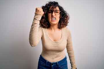 Young beautiful curly arab woman wearing casual t-shirt and glasses over white background angry and mad raising fist frustrated and furious while shouting with anger. Rage and aggressive concept.