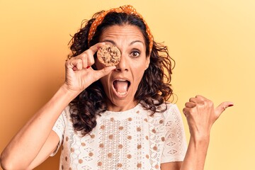 Wall Mural - Middle age beautiful woman holding chocolate cookie over eye pointing thumb up to the side smiling happy with open mouth