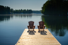 Two Adirondack Chairs On A Wooden Dock Overlooking A Calm Lake In Ontario Cottage Country. Retirement Planning.