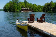 Two Adirondack Chairs On A Wooden Dock Facing A Calm Lake In Muskoka, Ontario Canada. A Yellow Canoe Is Tied To The Dock. A Cottage Nestled Between Trees Is Visible Across The Water.