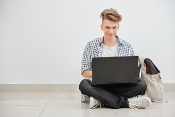 smiling high school student sitting on the floor and working or playing on laptop