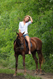Fototapeta Konie - Beautiful young girl with blond hair in a jacket with a shorts, smiling and riding her horse. Equestrian, lifestyle.