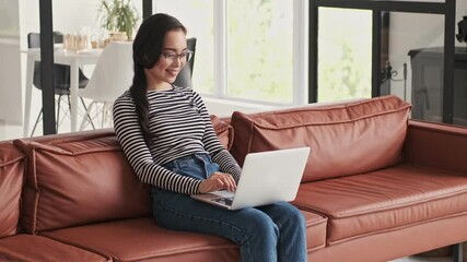 Wall Mural - Happy pretty asian woman in eyeglasses using laptop computer then having rest while sitting on sofa at home