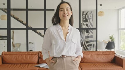 Wall Mural - Cheerful elegant pretty asian woman posing with arms in pockets and looking at the camera at home