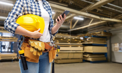 repair, construction and building concept - female worker with smartphone, helmet and working tools on belt over workshop at woodworking factory background