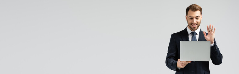 smiling  handsome businessman in suit having video chat on laptop isolated on grey, panoramic shot