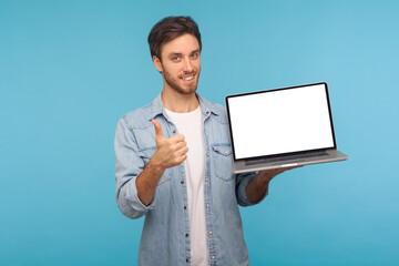 Portrait of satisfied smiling handsome man in worker denim shirt showing thumbs up and holding laptop screen with empty display, mock up for commercial image. studio shot isolated on blue background