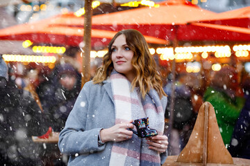 Wall Mural - Beautiful young woman drinking hot punch, mulled wine on German Christmas market. Happy girl in winter clothes with lights on background on winter snow day in Berlin, Germany.