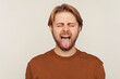 Portrait of funny optimistic man with beard wearing sweatshirt behaving childish naughty, showing tongue out and making faces, disobedient goofy grimace. indoor studio shot isolated on gray background