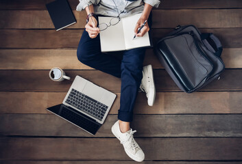 Man holding pencil writing notepad paper learning and research course tutoring education online on laptop with the coffee cup, briefcase, book on my desktop. Are sitting on the wooden floor. Top view