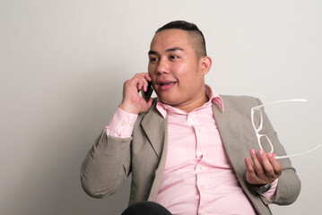 Wall Mural - Happy young overweight Asian businessman in suit sitting and talking on the phone