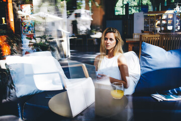 Pretty woman holding cup of coffee and dreaming about something while waiting for laptop computer restarts, young female skilled freelancer thinking about new ideas while sitting with net-book in cafe