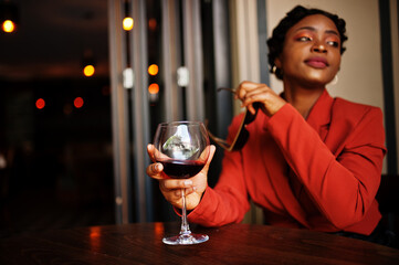 Portrait of african american woman, retro hairstyle with eyeglasses, wear orange jacket posing at restaurant with glass of wine.