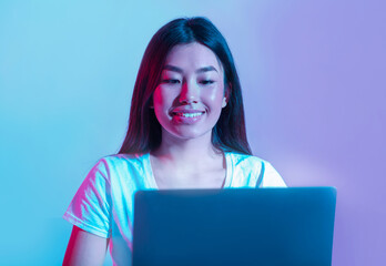 Work or play at computer. Smiling asian girl looking at laptop in neon