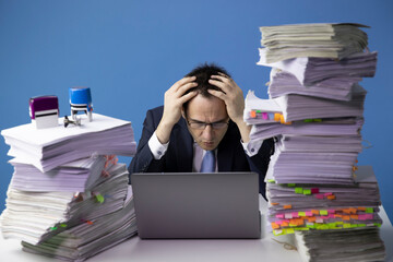 Office clerk works behind laptop, looks amazed at screen and takes up his head. Unorganized office work. Paperwork document stack, disarray in files on table. Routine paperwork. Overworked concept.