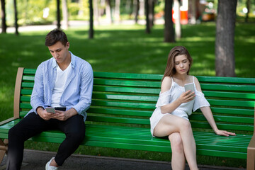 Wall Mural - Gadget addiction and relationship problems. Young couple with smartphones sitting on bench at park, ignoring each other
