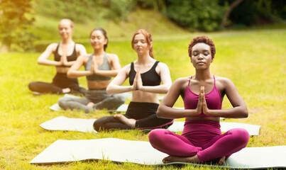 Wall Mural - Diverse millennial women meditating in lotus pose and doing namaste gesture on their outdoor yoga class, empty space