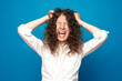 © Yulia - Stressful young curly haired female made terrible mistake, keeps both hands on head, eyes closed, opens mouth widely and yells angrily, wears casual cotton shirt, models over blue studio background..