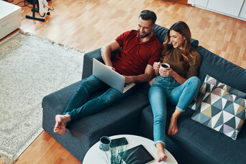 Sticker - Top view of carefree young couple in casual clothing bonding together and using laptop while resting on the sofa indoors