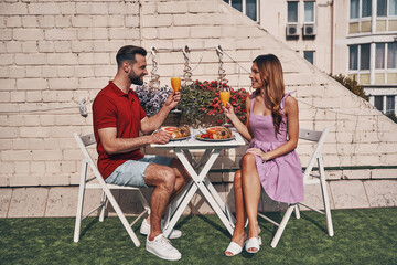 Sticker - Carefree young couple in casual clothing drinking juice and smiling while sitting on the rooftop patio outdoors