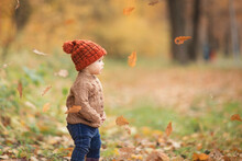 Cute Baby In Autumn Clothes Looks To The Right. Child In Knitted Hats And Jacket In Cool Weather. Girl Looks At Fall Of Oak Leaves.