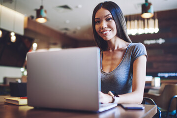 Portrait of happy female student resting in comfortable cafeteria with modern laptop computer for e learning, successful hipster girl using netbook for communicate with friends via application