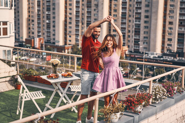 Canvas Print - Playful young couple in casual clothing dancing and smiling while having romantic dinner on the rooftop patio outdoors