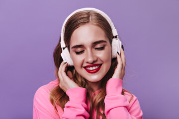 Good looking woman in pink hoodie listening to music in headphones on purple background. Happy girl in sweatshirt enjoying song on isolated