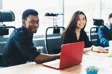 Portrait of positive young man in formal wear laughing at camera while discussing working strategy with male and female colleagues.Cheerful dark skinned employee sitting at meeting table with laptop