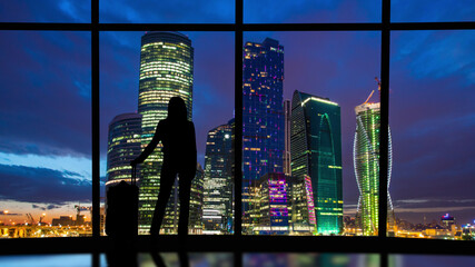 The woman with suitcase stands near the windows in the business office building