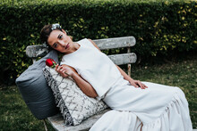 Attractive Young Lady In White Dress And With Red Rose Leaning On Soft Pillows And Looking Away While Resting On Shabby Bench On Wedding Day In Park