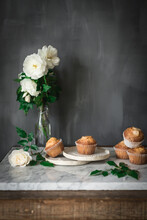 Still Life Of Tasty Yogurt Cupcakes On Round Plates In Arrangement With Green Leaves And Aromatic White Roses In Glass Vase On Marble Table Against Gray Wall In Kitchen