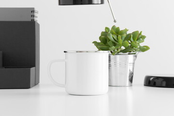 Enamel mug mockup with a lamp, succulent plant and workspace accessories on a white table.