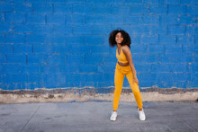 Delighted Ethnic Female In Yellow Clothes In Moment Of Dancing On Background Of Vivid Blue Wall On Street
