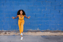 Calm African American Female With Afro Hairstyle And In Yellow Outfit Leaning On Blue Brick Wall In City And Stretching Arms At Wall