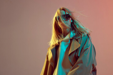 Stylish young girl, cool posing in a hoodie, sunglasses and cloak with developing hair, on a neon background.