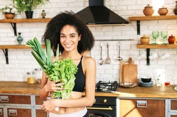 Healthy lifestyle concept. Young sporty woman in the kitchen with armful of fresh greenery and salads smiling