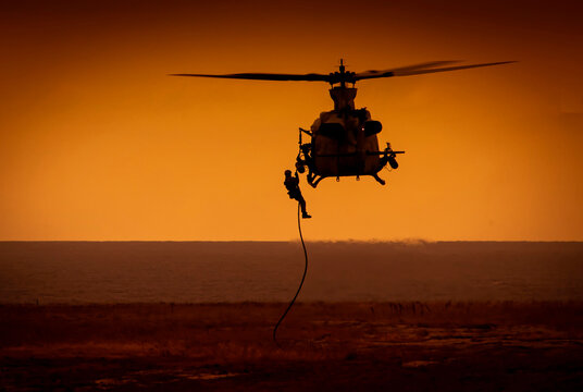 Shihoette of helicopter lowering a soldier by rope to the ground with ocean in view at sunset