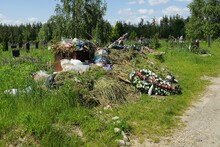 A Large Pile Of Garbage With Old Funeral Wreaths And Dry Vegetation In The Green Grass Among The Graves And Monuments In The Cemetery
