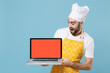 Shocked young bearded male chef or cook baker man in apron white t-shirt toque chefs hat isolated on blue wall background studio. Cooking food concept. Hold laptop pc computer with blank empty screen.