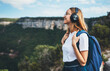 © A_B_C - positive young traveler girl with headphones and backpack stands on top of mountain, active tourist woman hiking enjoys beautiful landscape and . fresh air high in mount,  empty space for text
