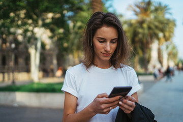 Wall Mural - pretty woman writes message on mobile smartphone while walking Park barcelona on a warm summer day, hipser girl listens to music on gadget searches for information using cell phone