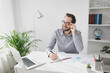 © ViDi Studio - Smiling young bearded business man in gray shirt glasses sit at desk work on laptop pc computer in light office on white wall background. Achievement business career concept Writing notes in notebook.