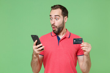 Wall Mural - Shocked young bearded man guy in casual red pink t-shirt posing isolated on green background studio portrait. People lifestyle concept. Mock up copy space. Using mobile phone, hold credit bank card.