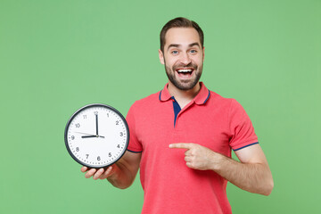 Excited young bearded man guy in casual red pink t-shirt posing isolated on green background studio portrait. People emotions lifestyle concept. Mock up copy space. Pointing index finger on clock.