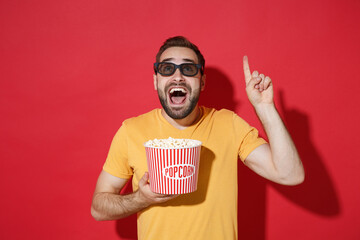 Wall Mural - Excited young man guy in casual yellow t-shirt 3d glasses isolated on red wall background. People in cinema, lifestyle concept. Watching movie film holding bucket of popcorn pointing index finger up.