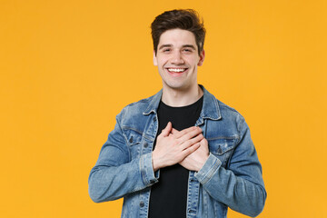 Smiling young man guy in casual denim jacket posing isolated on yellow background studio portrait. People sincere emotions lifestyle concept. Mock up copy space. Holding hands folded on chest, heart.