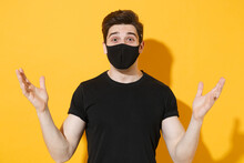 Shocked Young Man Guy In Black T-shirt Face Mask Isolated On Yellow Wall Background Studio Portrait. Epidemic Pandemic Spreading Coronavirus 2019-ncov Sars Covid-19 Flu Virus Concept. Spreading Hands.