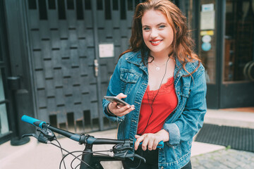 Wall Mural - Red curled long hair caucasian teen girl on the city street walking with bicycle using the smartphone with earphones fashion portrait. Natural people beauty urban life concept image.