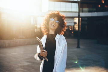 Curly haired businesswoman wearing eyeglasses and smiling at camera is posing in a sunny day with a laptop